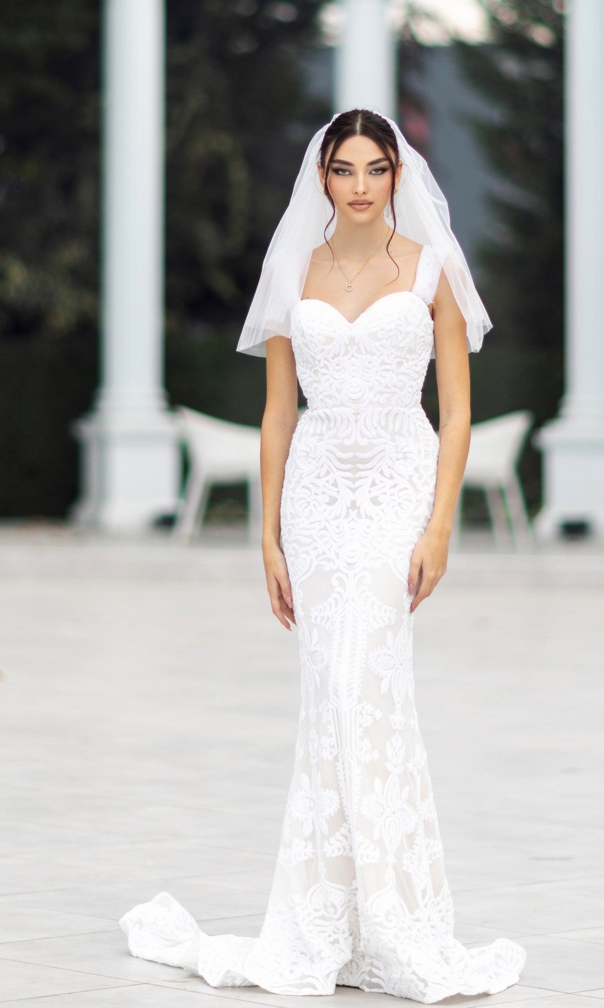 A woman wears the Off-Shoulder White Bridal Gown: Tina Holly TW103 and a veil, standing outdoors on a paved area with white columns and chairs in the background.